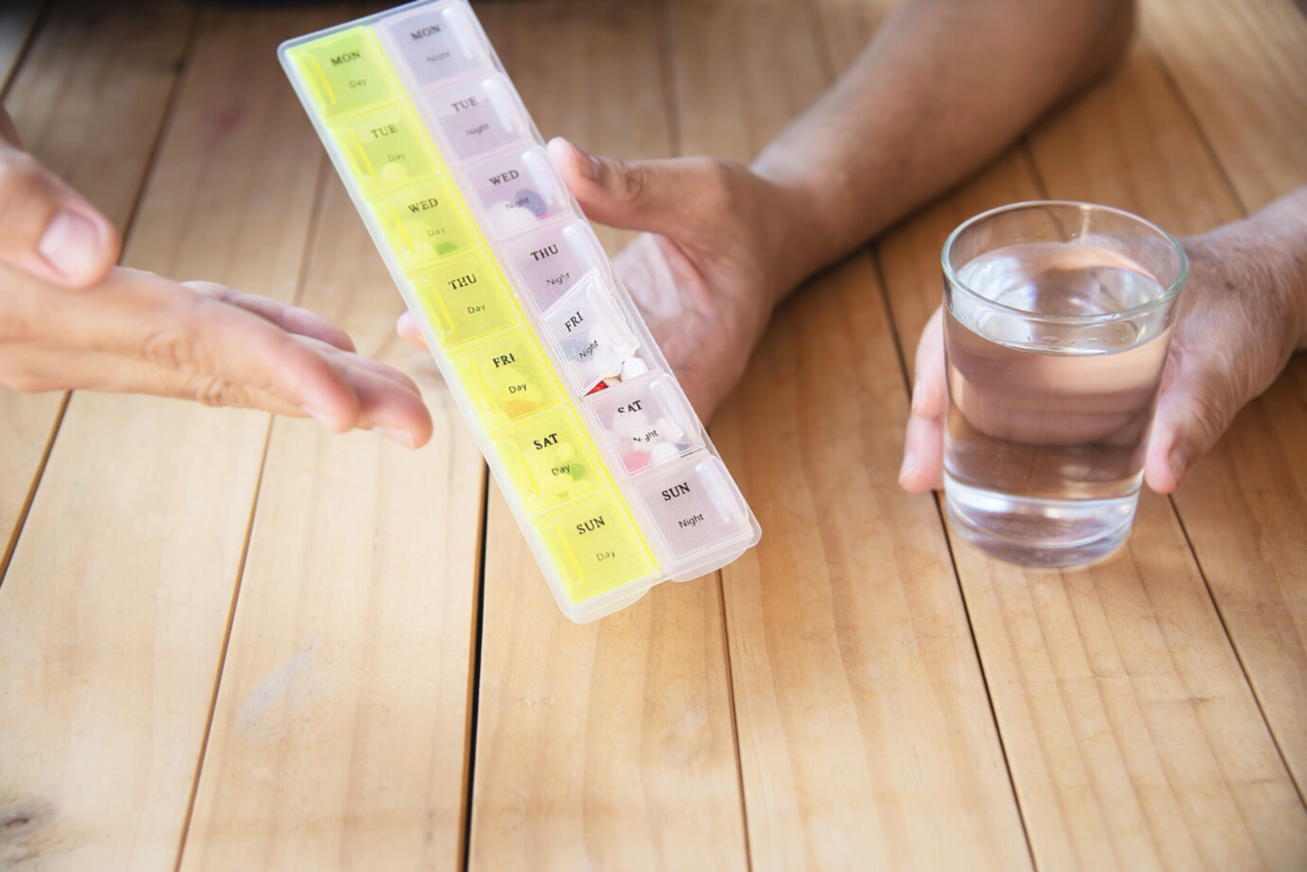 Two hands hold a pill organizer labeled with days of the week and a glass of water on a wooden table, suggesting a medication routine.