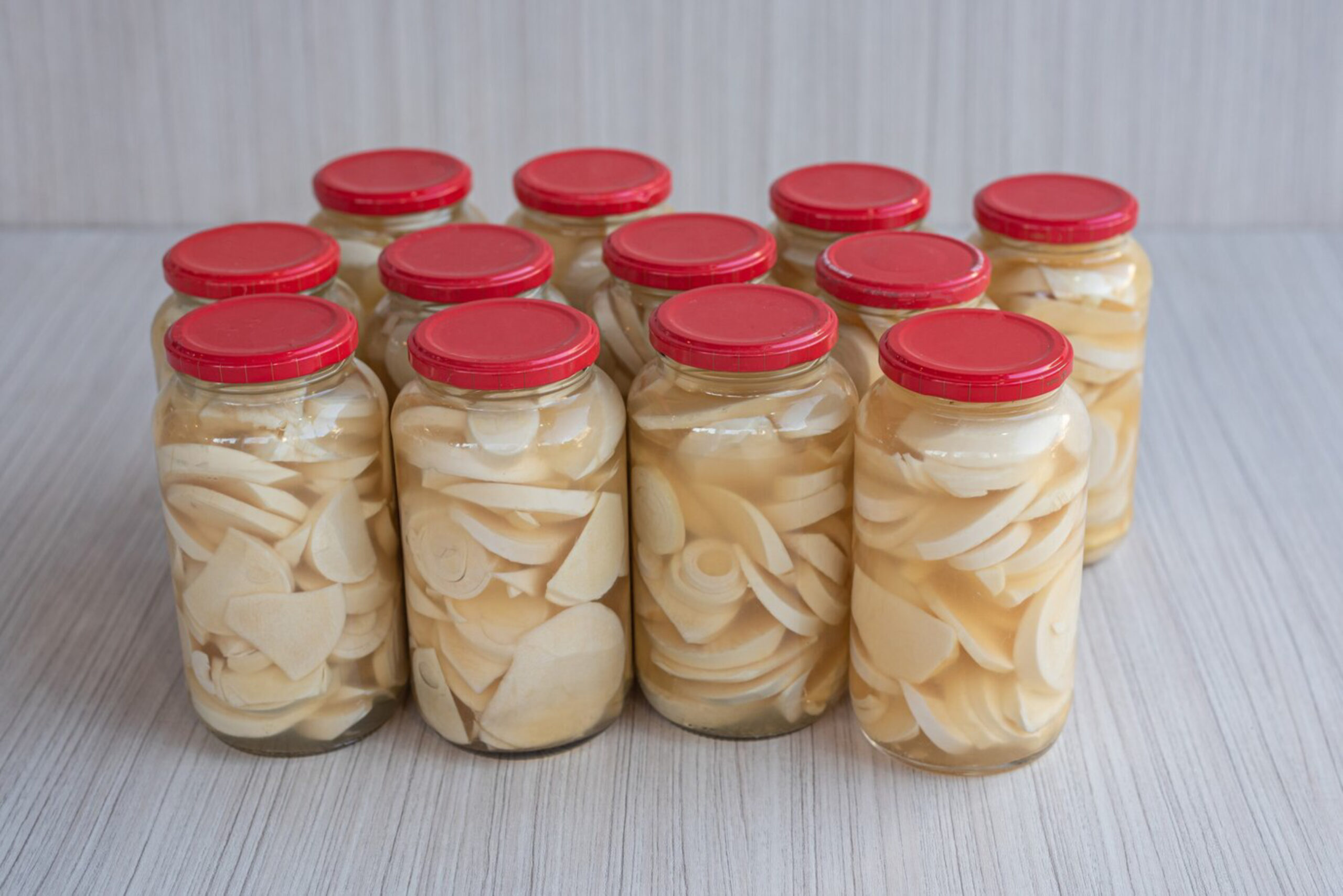 Ten jars filled with sliced pickled vegetables are arranged in a neat group on a light wooden surface. Each jar has a vibrant red lid, creating a fresh and organized look.