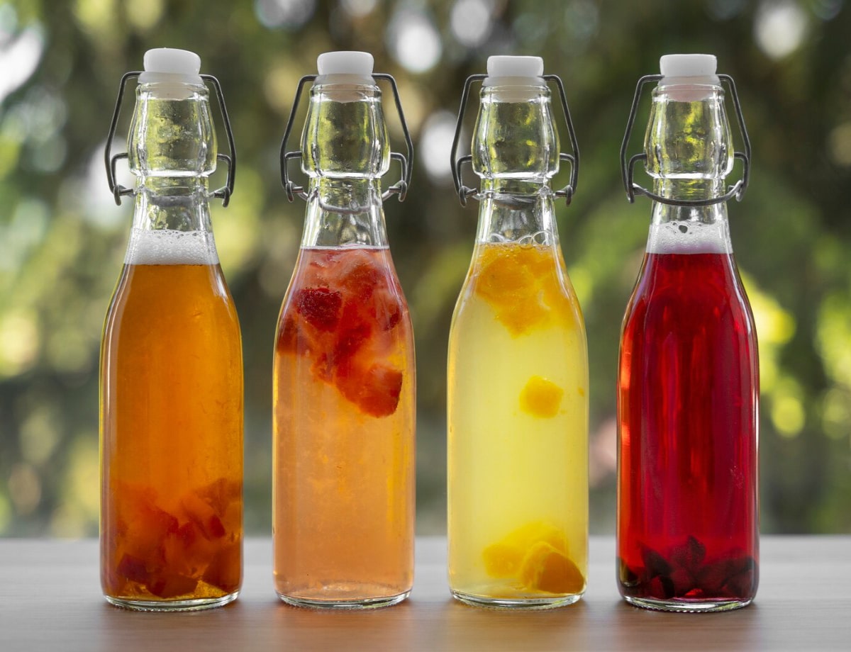 Four glass bottles filled with colorful, fruity beverages, featuring orange, pink, yellow, and red liquids against a blurred green backdrop.