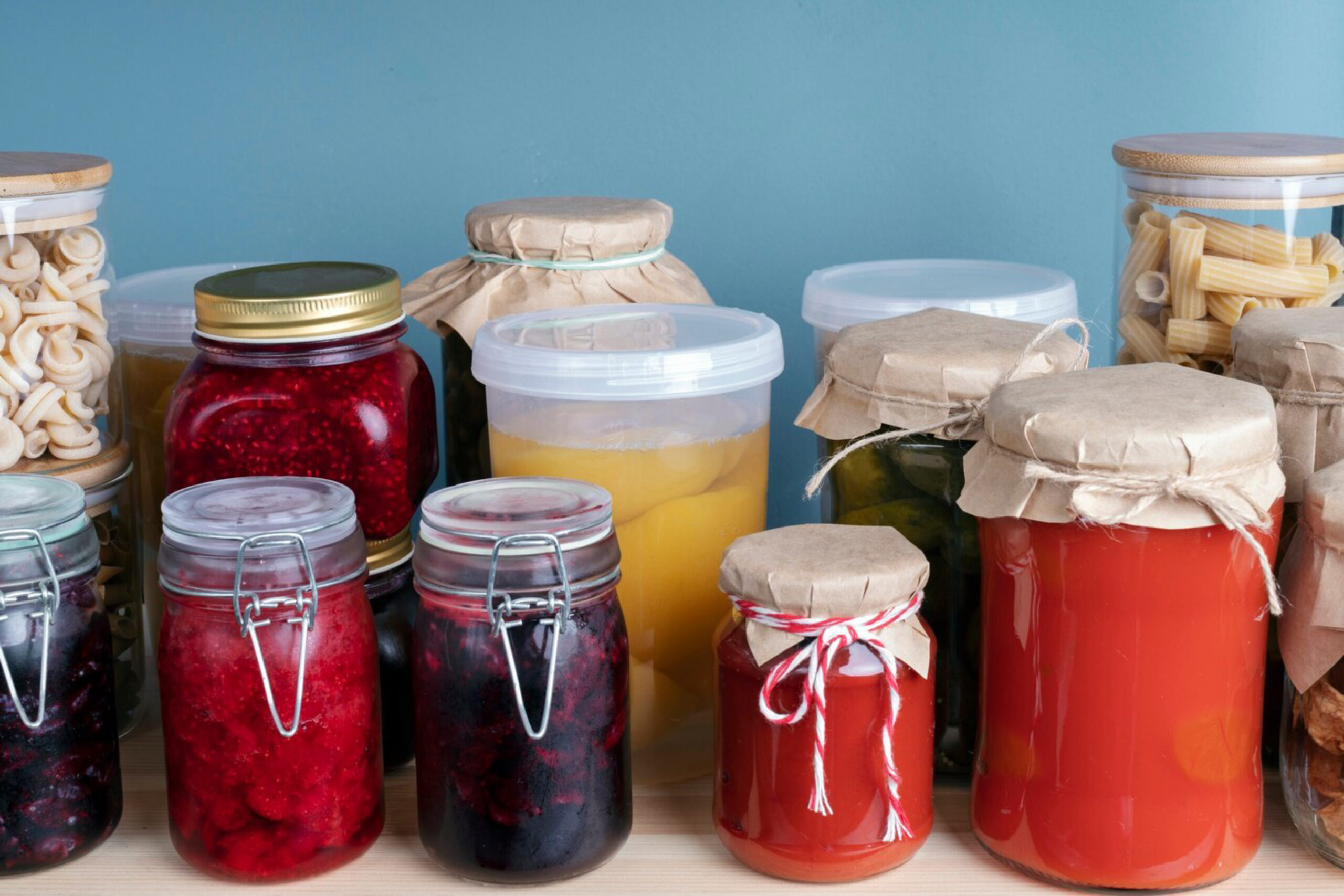 Various jars and containers filled with preserved foods, including pasta, jams, and pickles, are arranged against a light blue background.