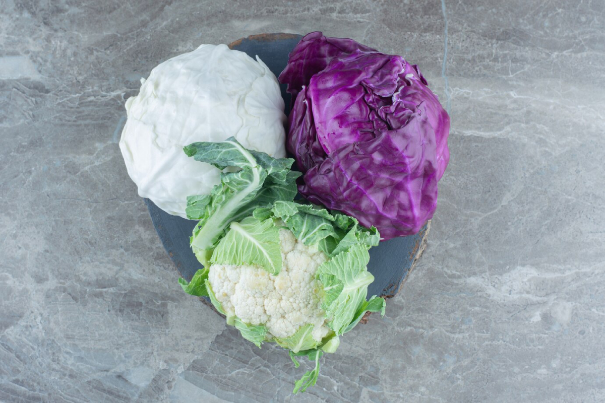 Three types of cabbage—white, purple, and cauliflower—arranged on a round wooden platter against a marble background.