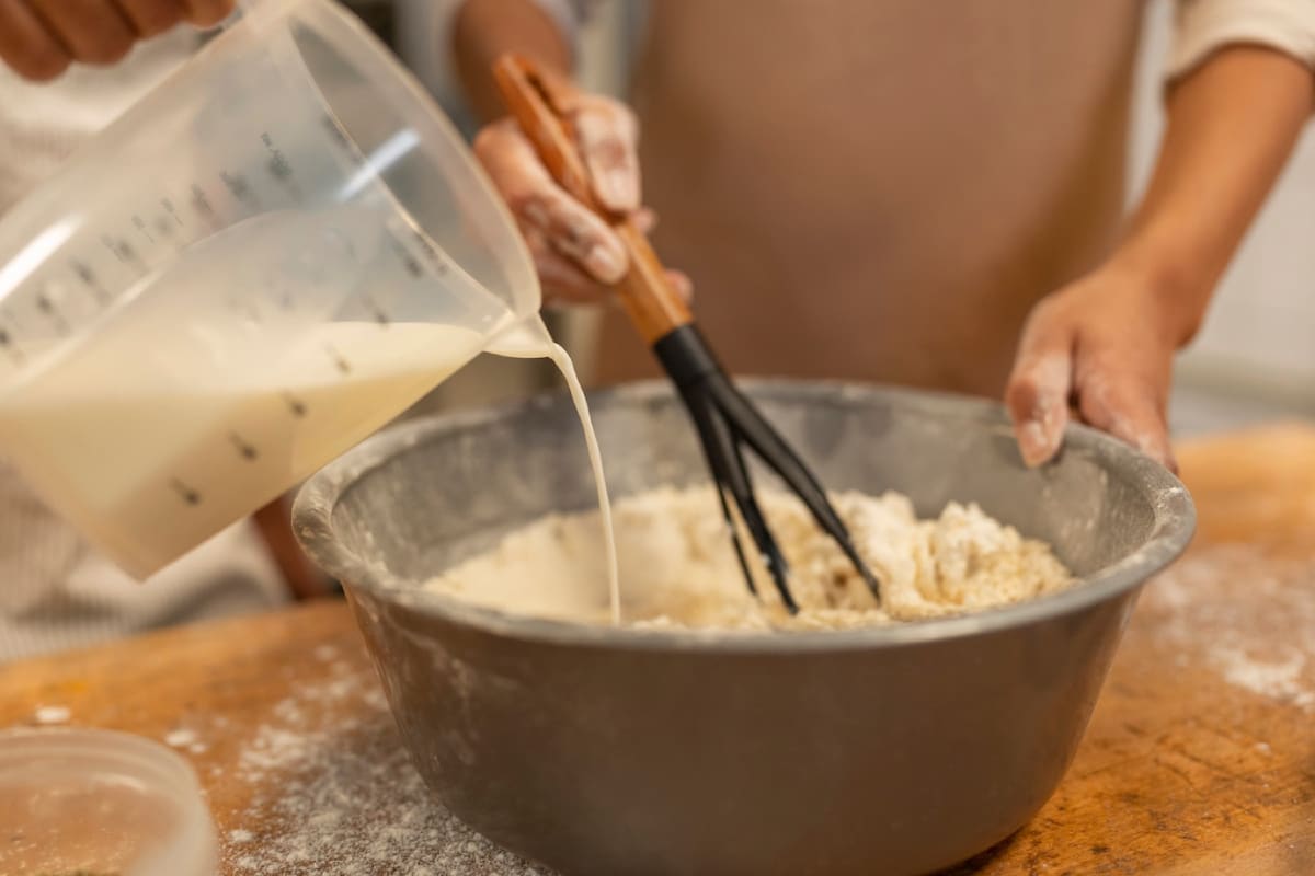 A person pours milk from a measuring cup into a mixing bowl filled with flour, while stirring with a whisk. Flour dust is visible on the table.