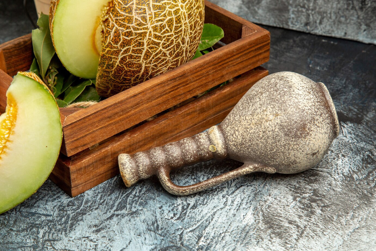A wooden crate with melon halves, leaves, and an ancient-looking ceramic jug on a textured surface.