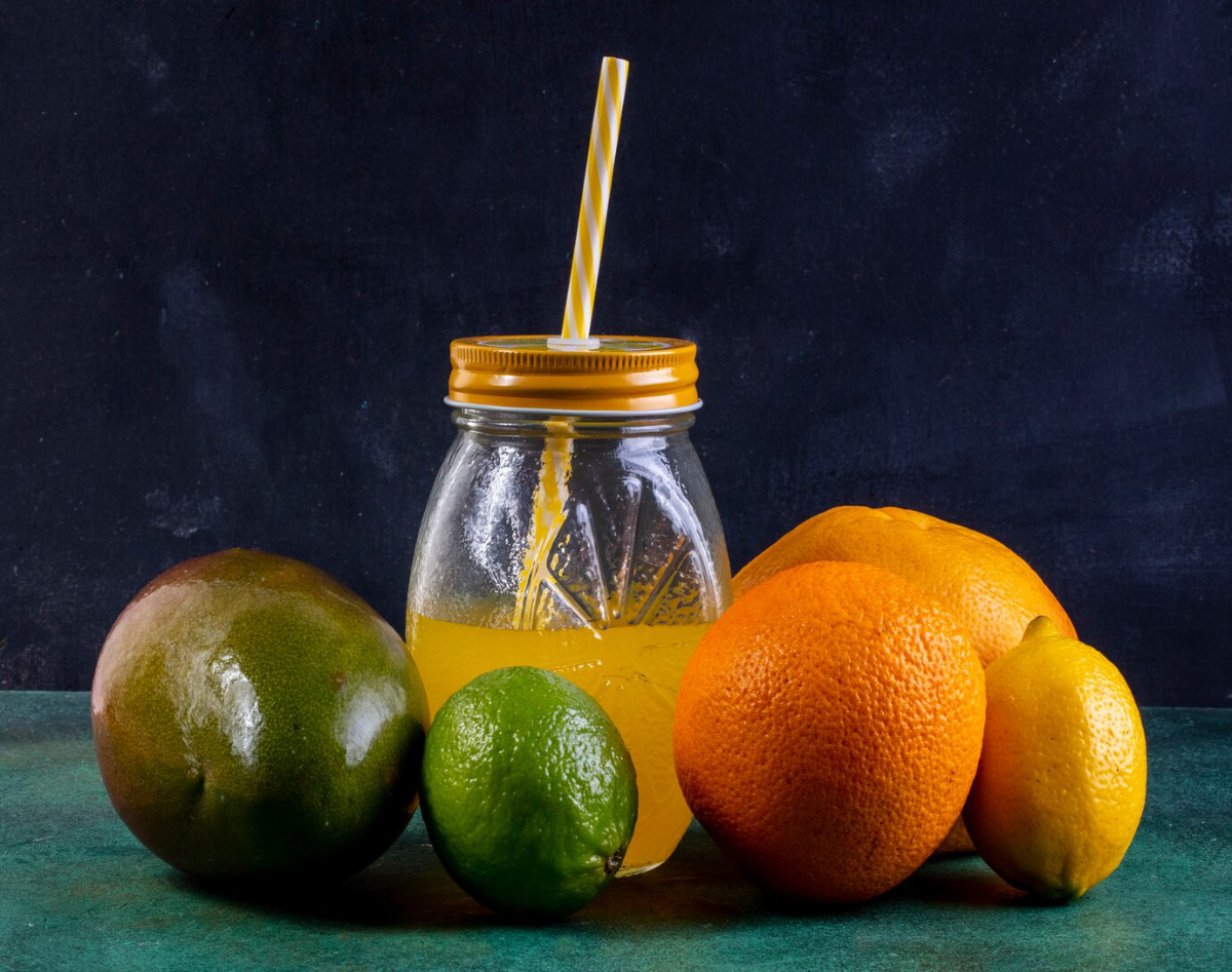 A jar of fresh juice with a striped straw, surrounded by a green mango, lime, orange, and lemon on a teal surface.