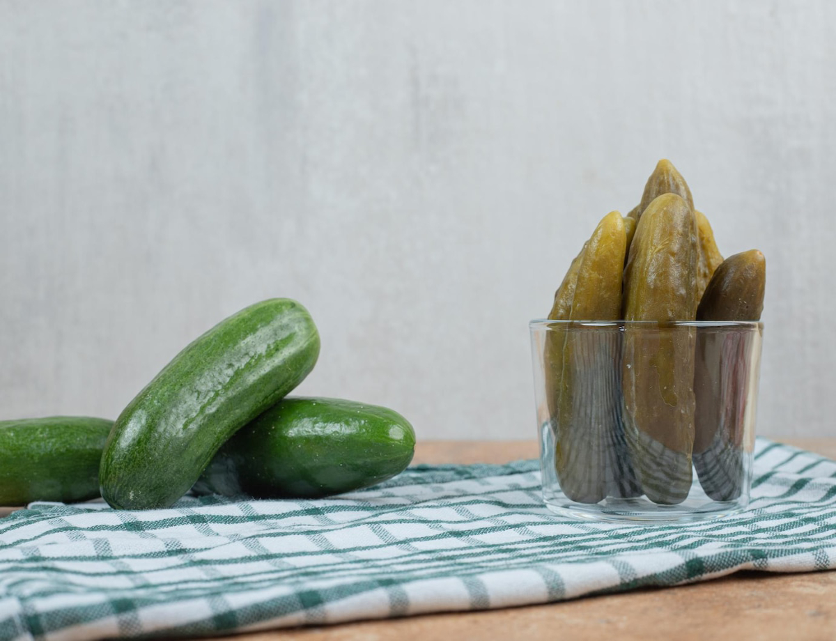 Three fresh green cucumbers rest on a checkered kitchen towel next to a glass filled with pickles.