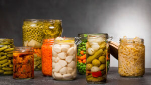 A variety of colorful pickled vegetables in glass jars, including carrots, cucumbers, olives, and sprouts, arranged on a dark surface.
