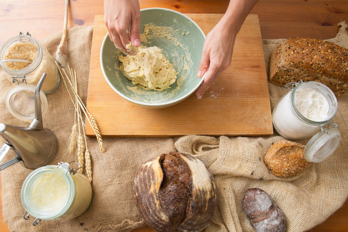 Hands mixing dough in a green bowl, surrounded by jars of ingredients and various types of bread on a rustic table.