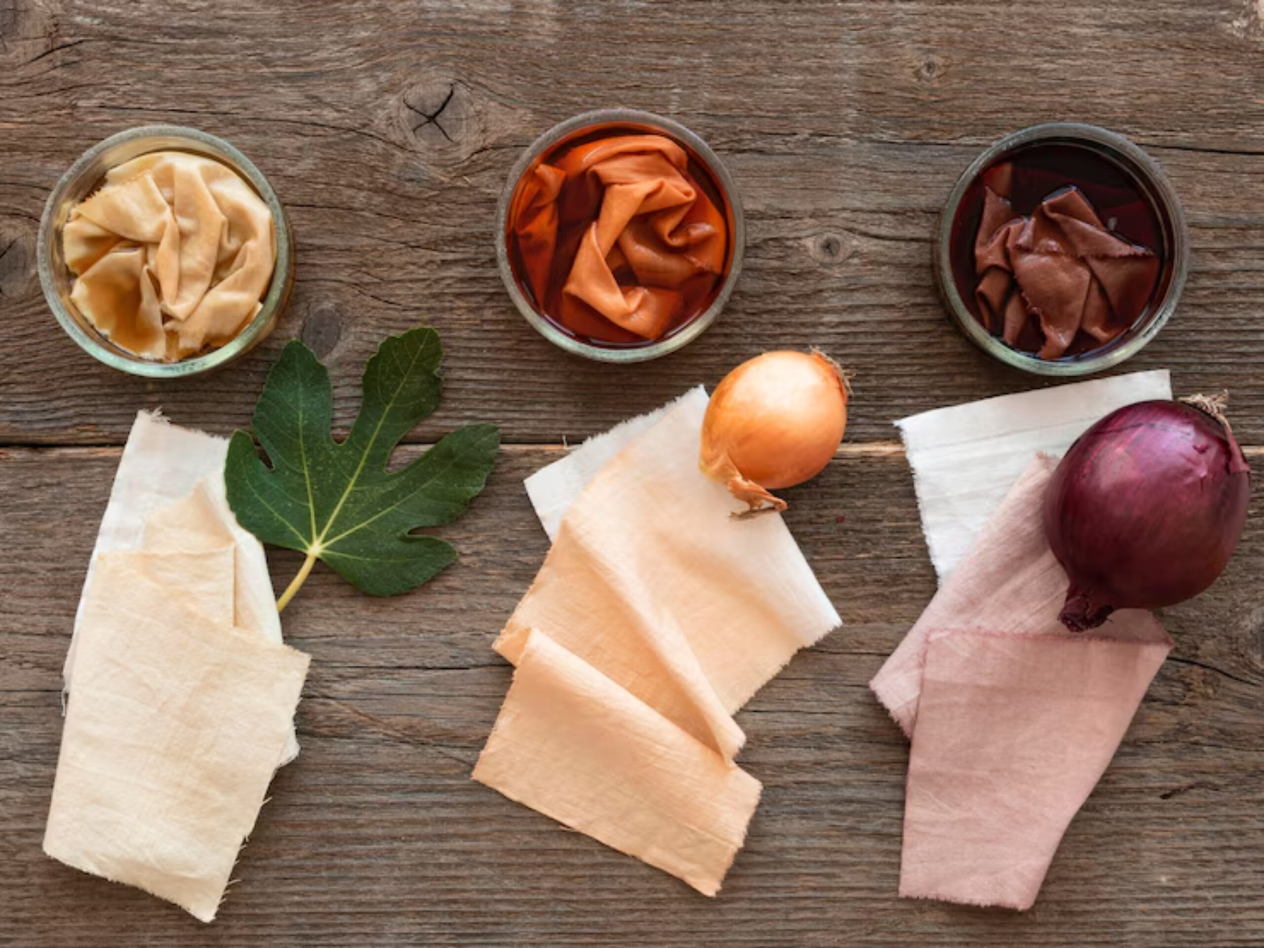 Three jars of dyed fabrics next to yellow and red onions, with a fig leaf on rustic wooden background. Natural dyeing process setup.