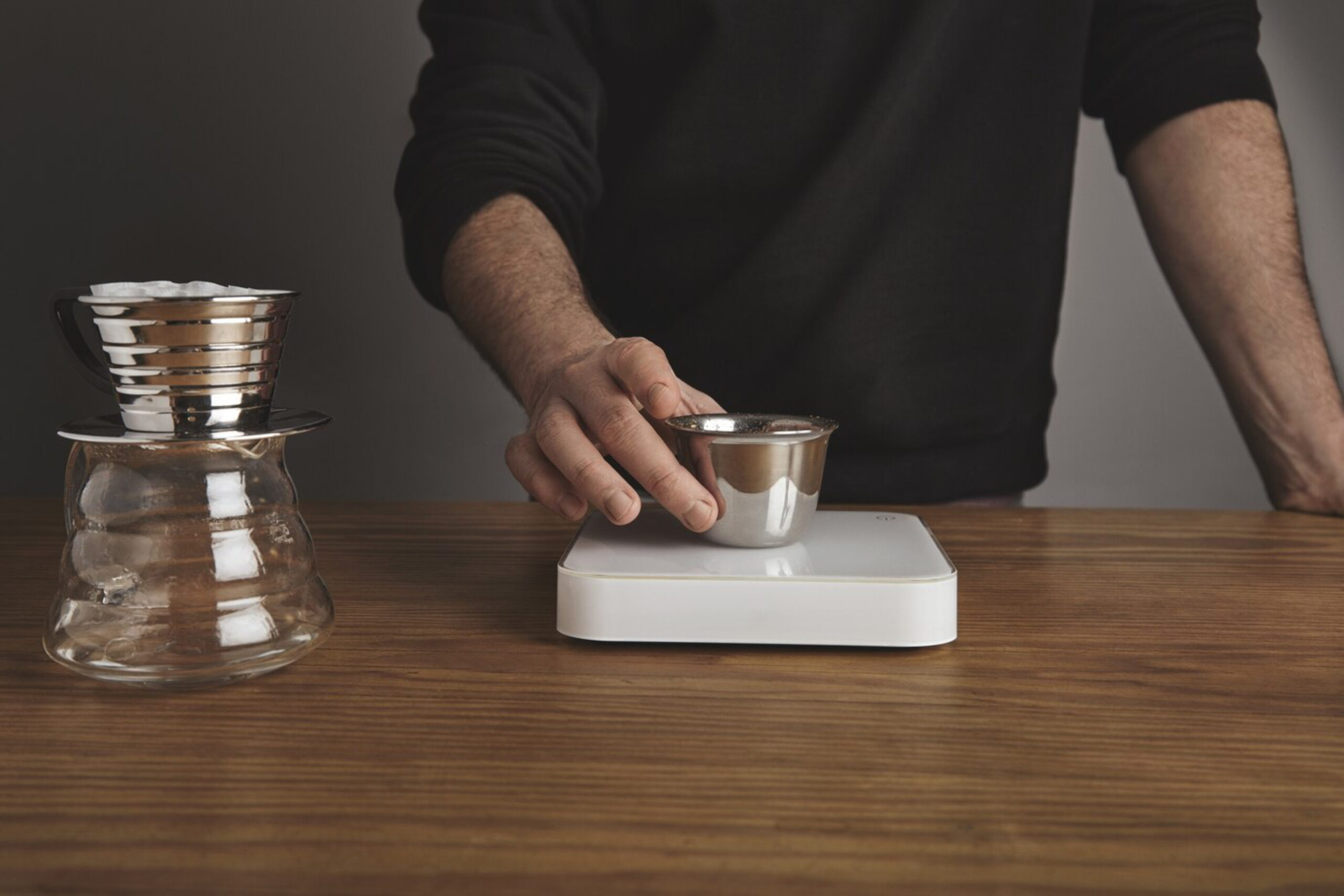 A person in a black sweater uses a scale to measure coffee grounds in a metal bowl on a wooden table, next to a pour-over coffee maker.