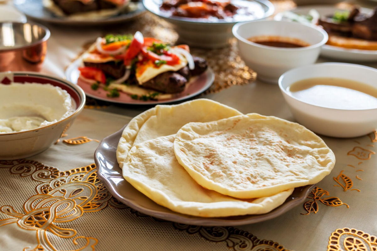 A plate of soft, round flatbreads sits on a decorative tablecloth, surrounded by various bowls of rich sauces and dishes.