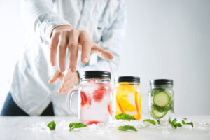 A hand reaches towards three jars of colorful, infused drinks surrounded by ice and mint leaves on a wooden table.