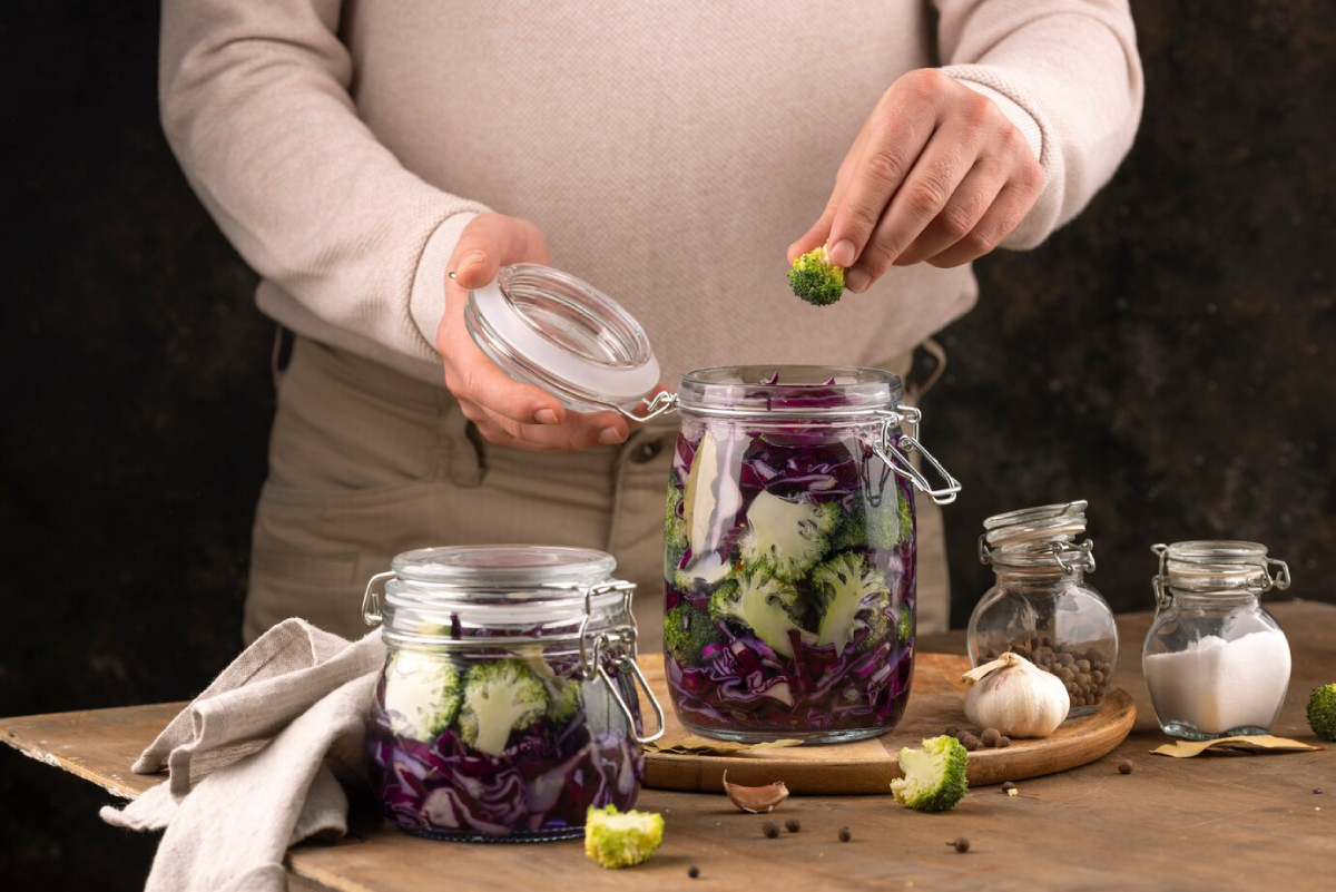 A person adds broccoli to jars of colorful pickled vegetables on a wooden table, surrounded by spices and garlic.
