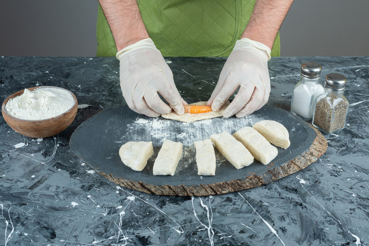 A person in gloves rolls dough with a piece of orange filling, surrounded by flour and seasonings on a rustic wooden board.
