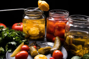 A collection of pickled vegetables in jars, surrounded by fresh tomatoes, cucumbers, and cilantro on a dark background.