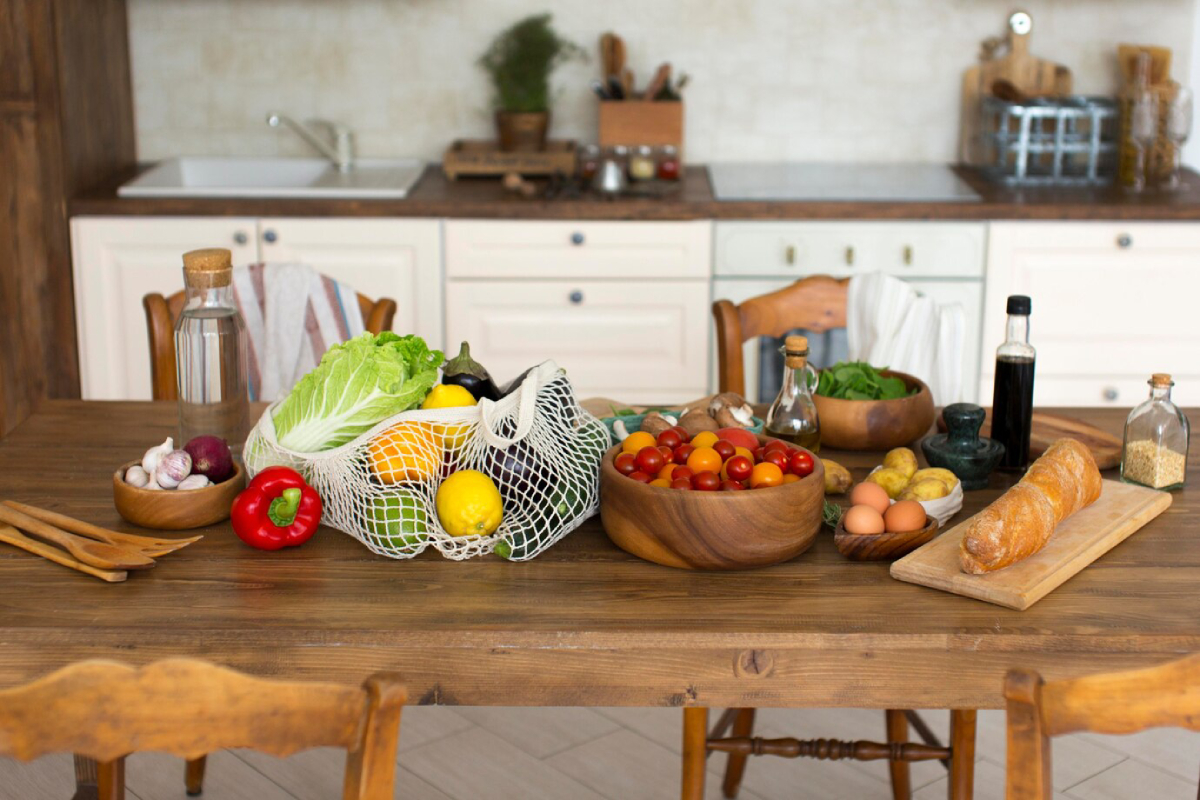 A wooden table displays an array of fresh vegetables, fruits, eggs, and bread, with olive oil and seasonings in a cozy kitchen setting.