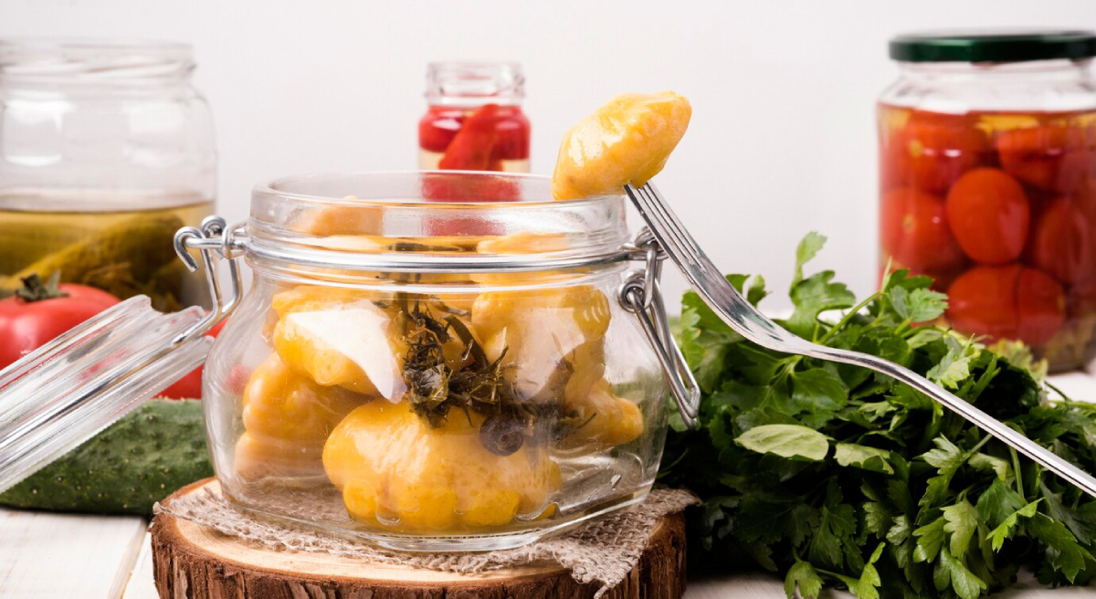A glass jar filled with yellow dumplings and herbs, with a fork poised over it; surrounded by fresh herbs, tomatoes, and pickles.