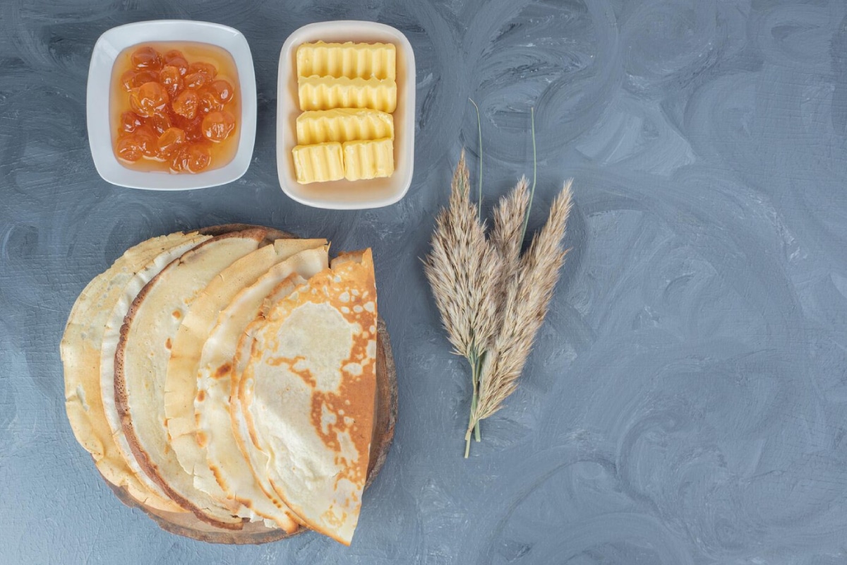 A wooden plate with stacked crepes, accompanied by butter, sweet apricot jam, and decorative dried grasses on a textured gray background.