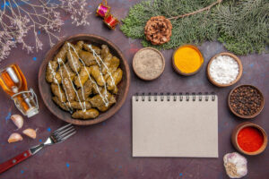 A wooden bowl of stuffed vine leaves drizzled with sauce, surrounded by spices, garlic, a notepad, and decorative elements.