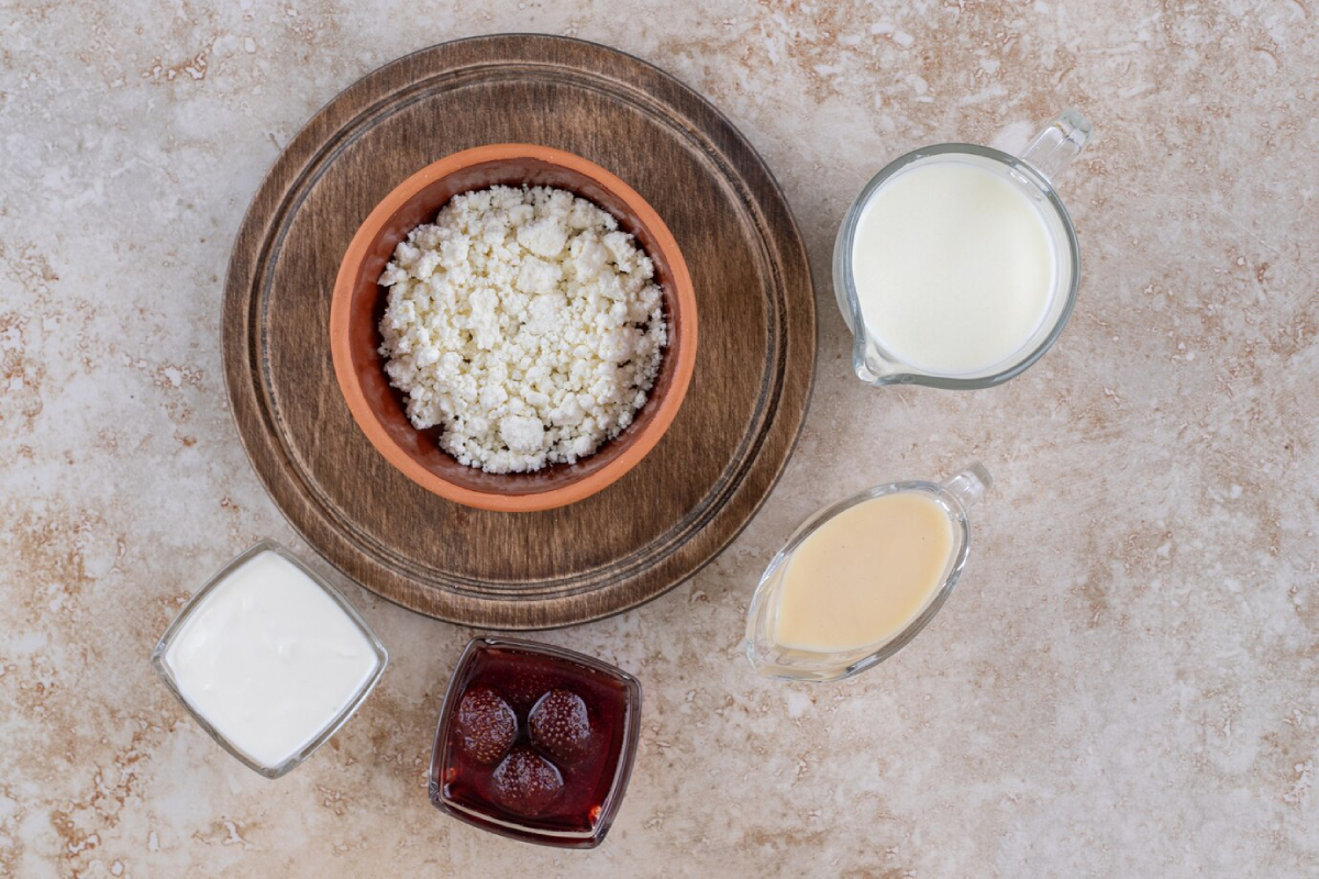 A terracotta bowl filled with cottage cheese, accompanied by cream, sweetened condensed milk, and strawberry jam on a wooden tray.