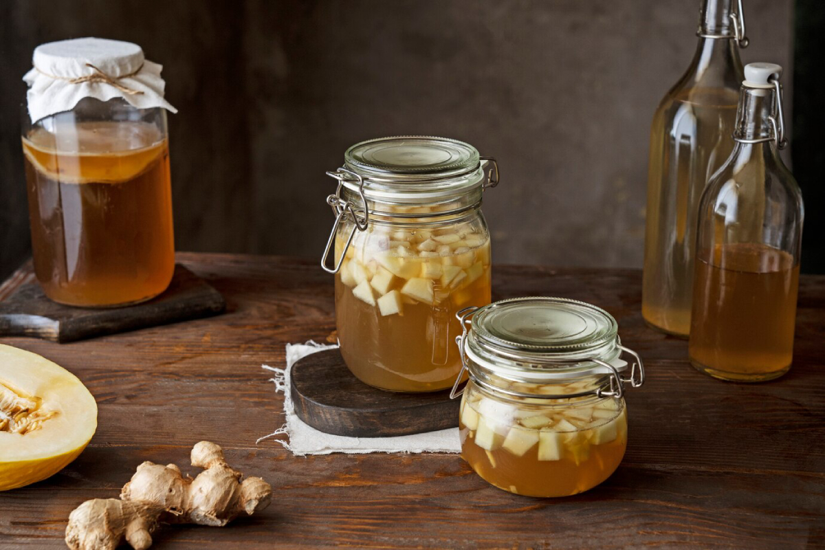 Two glass jars with pickled ginger and diced fruit, alongside a larger jar and two bottles of liquid, on a rustic wooden table.