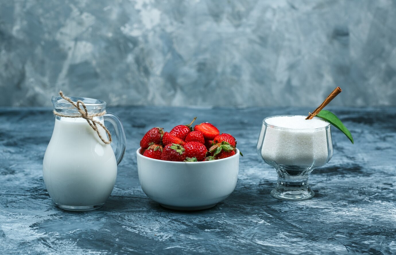 A glass pitcher of milk, a bowl of fresh strawberries, and a glass dish of sugar with a cinnamon stick and leaf on a textured blue background.
