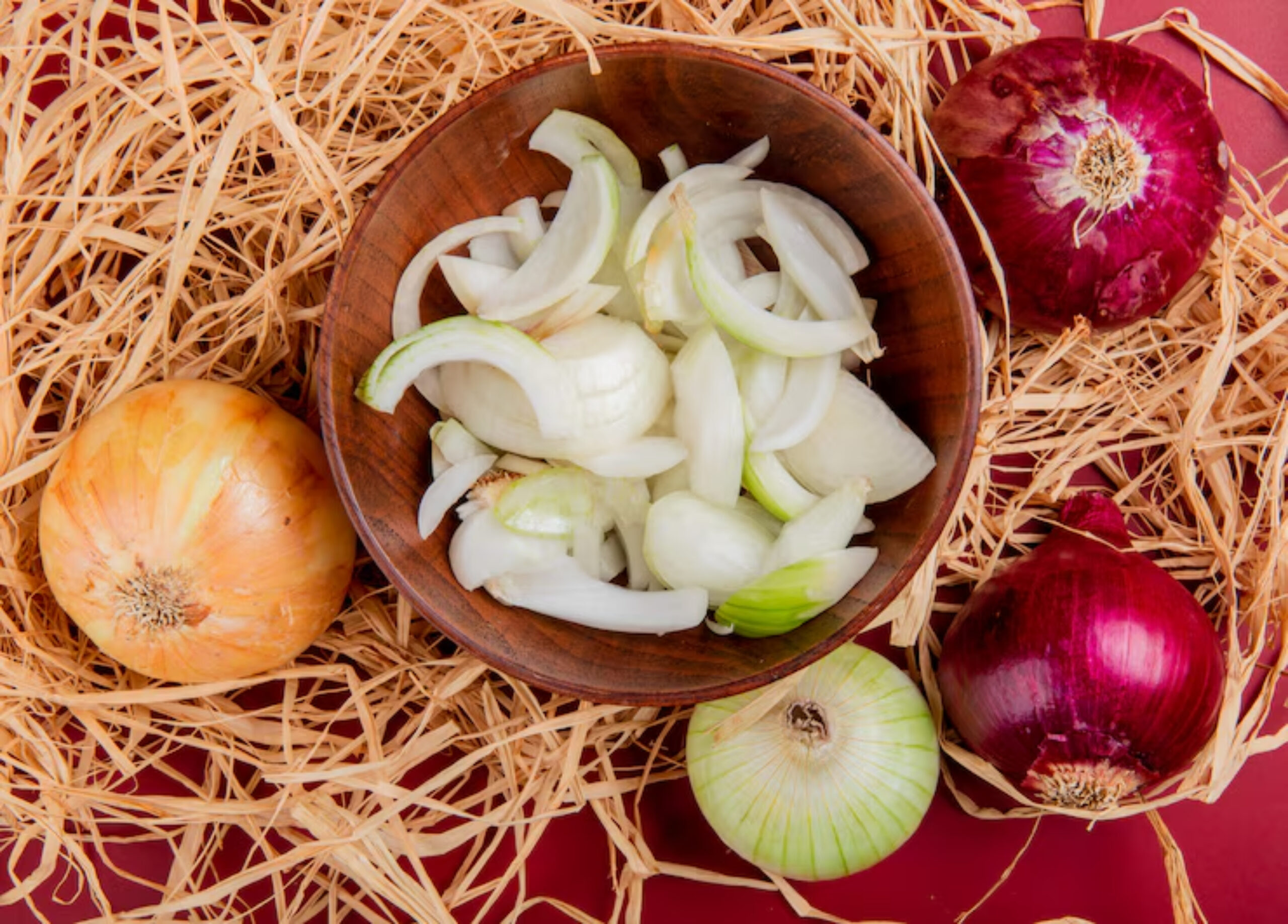 Wooden bowl of sliced white onions on straw, surrounded by whole yellow, white, and red onions, set against a red background. Rustic and fresh.