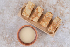 Four golden-brown crepes neatly arranged on a wooden board next to a small bowl of creamy sauce.