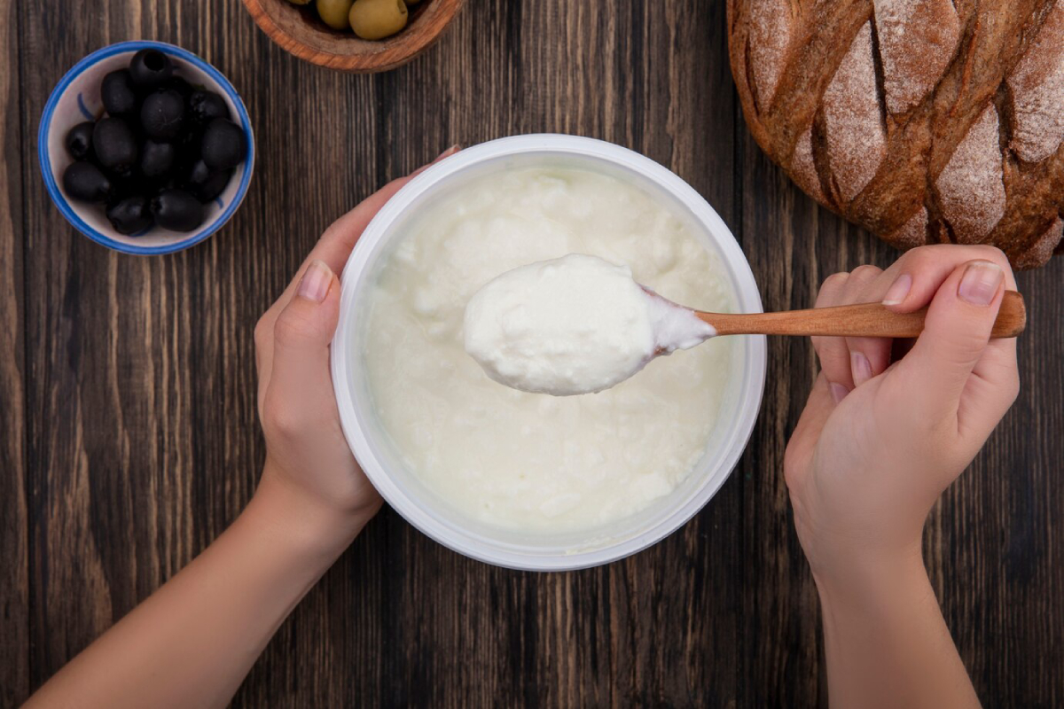 A hand holding a wooden spoon scoops from a container of yogurt, with bread and bowls of olives in the background.