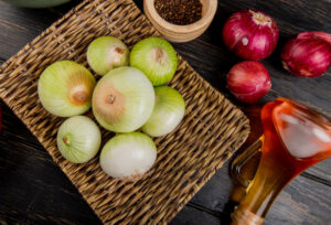 Wicker basket with white onions on a wooden surface. Nearby are red onions, whole peppercorns, and a glass jug of amber liquid, creating a rustic kitchen scene.