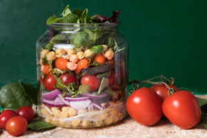 A glass jar filled with layers of colorful vegetables, including tomatoes, chickpeas, and greens, on a textured surface.