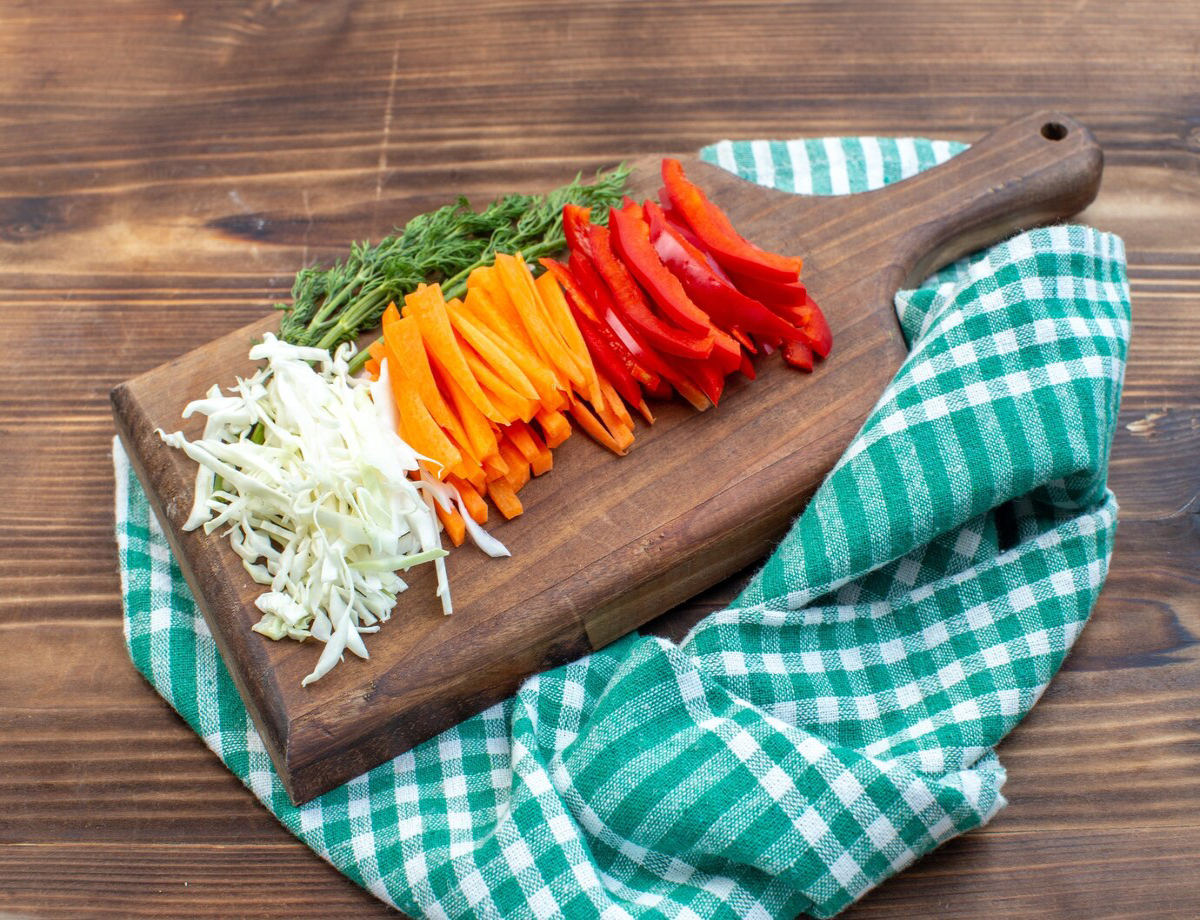 Colorful sliced vegetables including cabbage, carrots, and red bell peppers, garnished with fresh thyme on a wooden cutting board.
