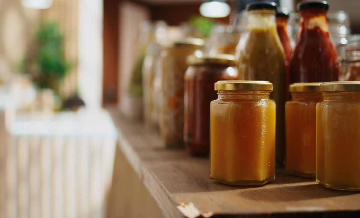 Glass jars filled with various sauces and preserves, placed neatly on a wooden shelf in a warm, inviting setting.