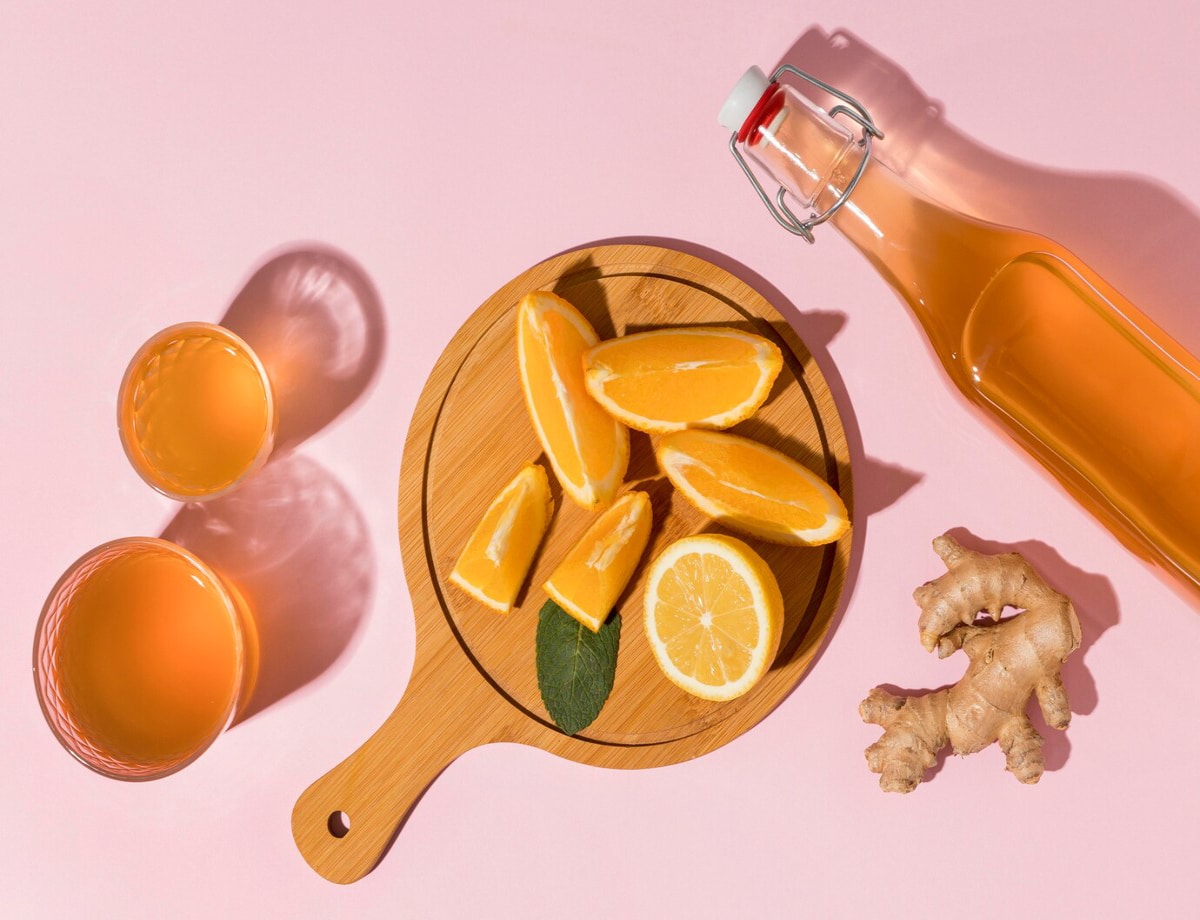 Sliced oranges and lemon on a wooden board with ginger root, alongside two glasses and a bottle of amber liquid, all on a pink background.