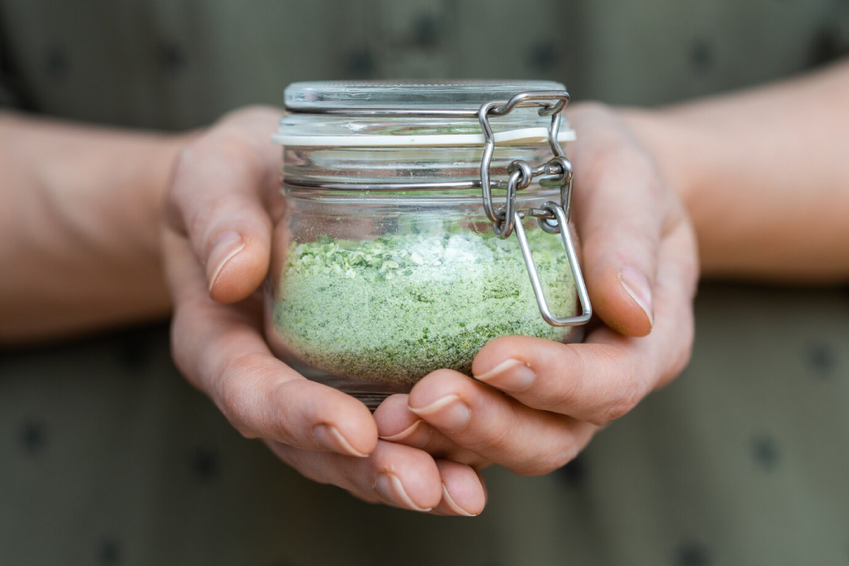 A person holding a small glass jar filled with green powder, showcasing a close-up of their hands and the jar's clasp.