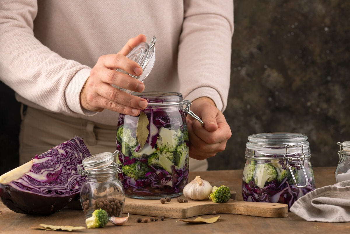 A person opening a jar of pickled vegetables, featuring red cabbage, broccoli, garlic, and spices on a wooden table.