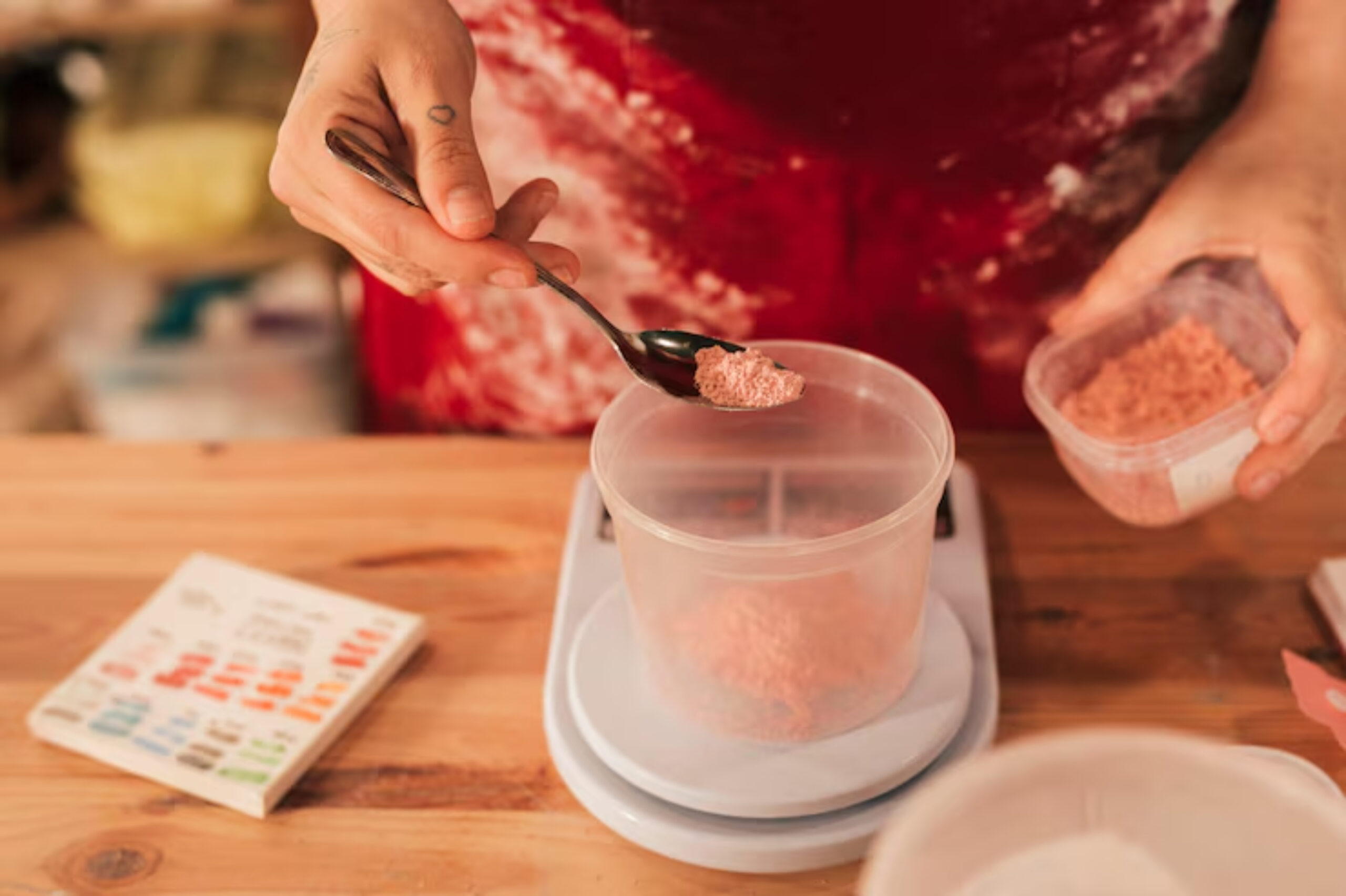 A person in a red apron, with a heart tattoo on their finger, is measuring pink powder into a plastic container on a digital scale, suggesting careful preparation.