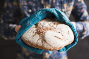 A person holds a freshly baked round loaf of bread, covered partially with a blue towel, revealing its crusty, flour-dusted surface.