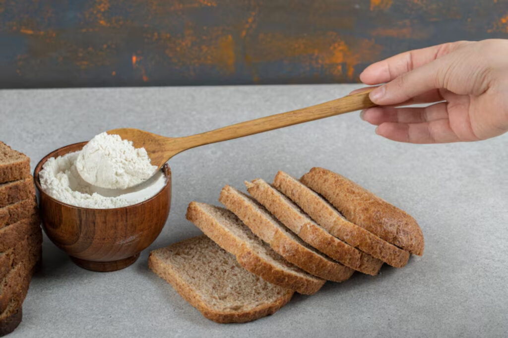 A hand holding a wooden spoon above a bowl of flour, next to slices of brown bread on a gray surface.