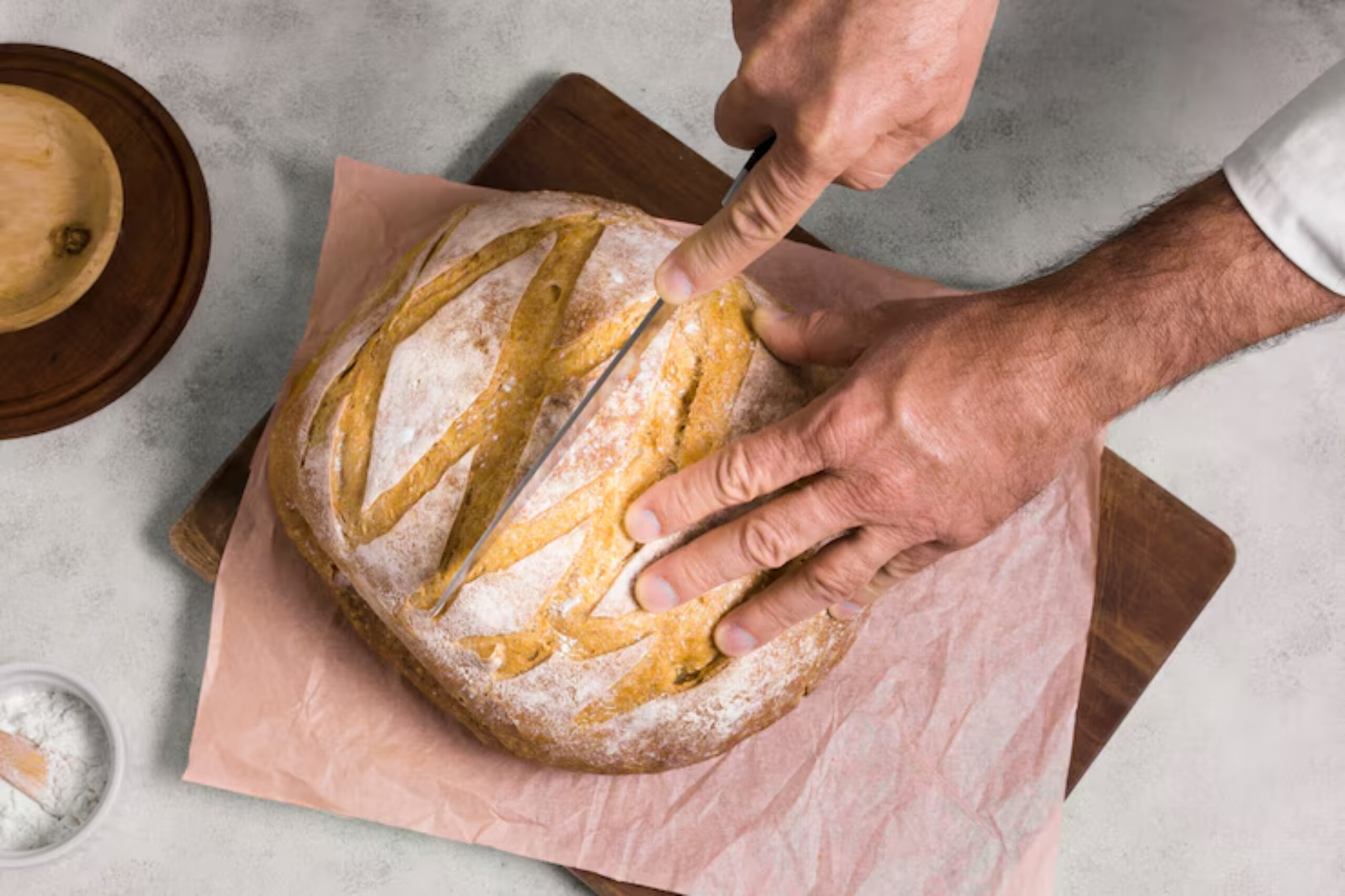 A hand slices a freshly baked loaf of bread on parchment paper, surrounded by a wooden bowl and a small dish of flour.