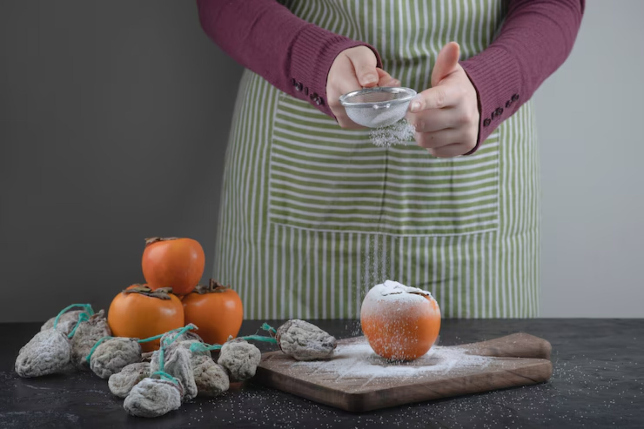 A person in a striped apron sprinkles powdered sugar over a persimmon on a cutting board, with dried fruits in the background.
