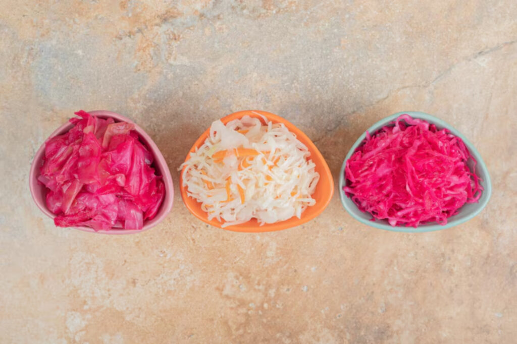 Three small bowls of fermented cabbage sit on a textured surface. The left has pink cabbage, the middle white, and the right vibrant magenta. Bright and colorful.