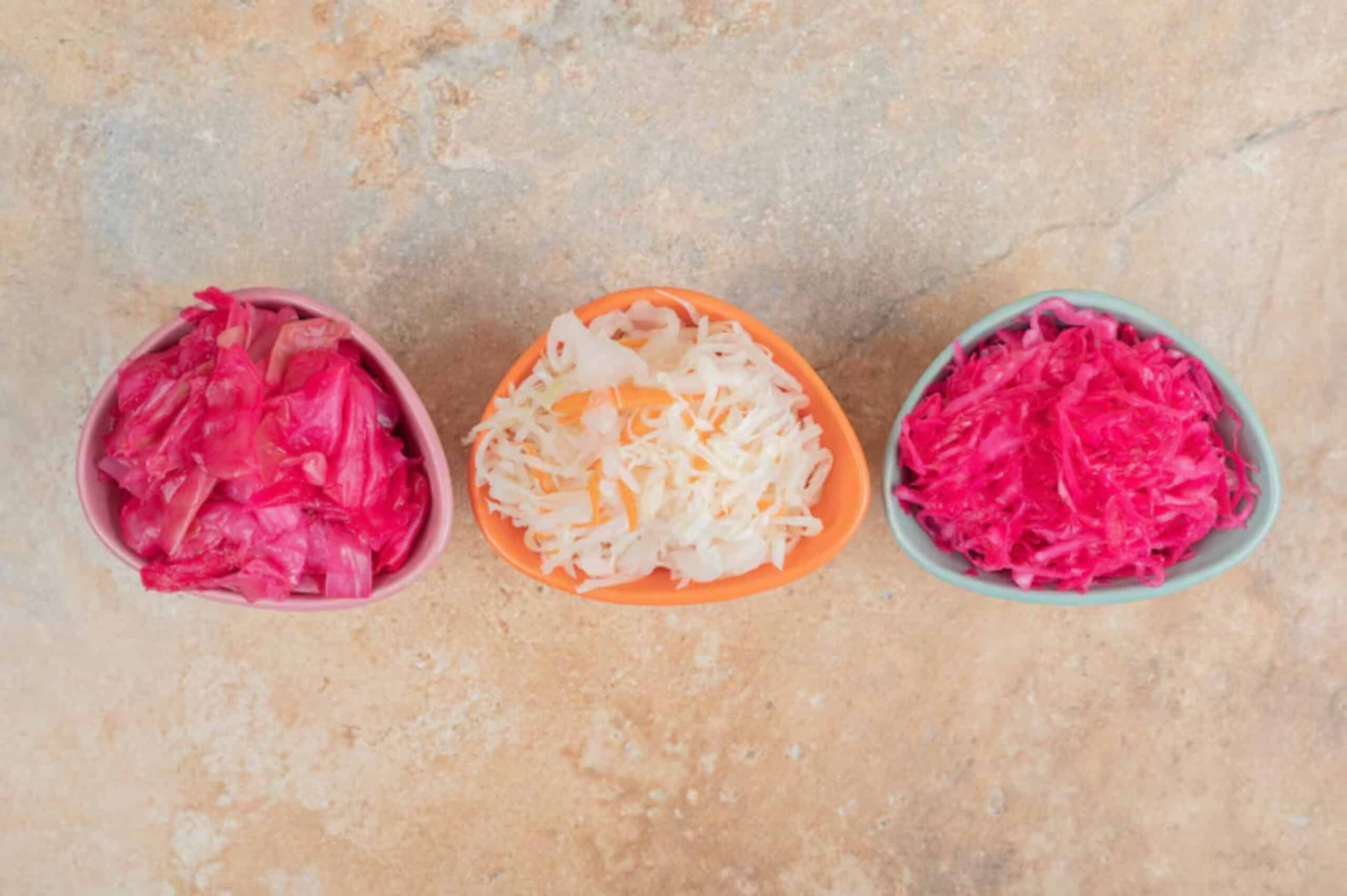Three small bowls of fermented cabbage sit on a textured surface. The left has pink cabbage, the middle white, and the right vibrant magenta. Bright and colorful.