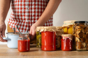 A person in a checkered shirt stands beside various jars of homemade preserves, including tomato sauce and pickles, on a wooden table.