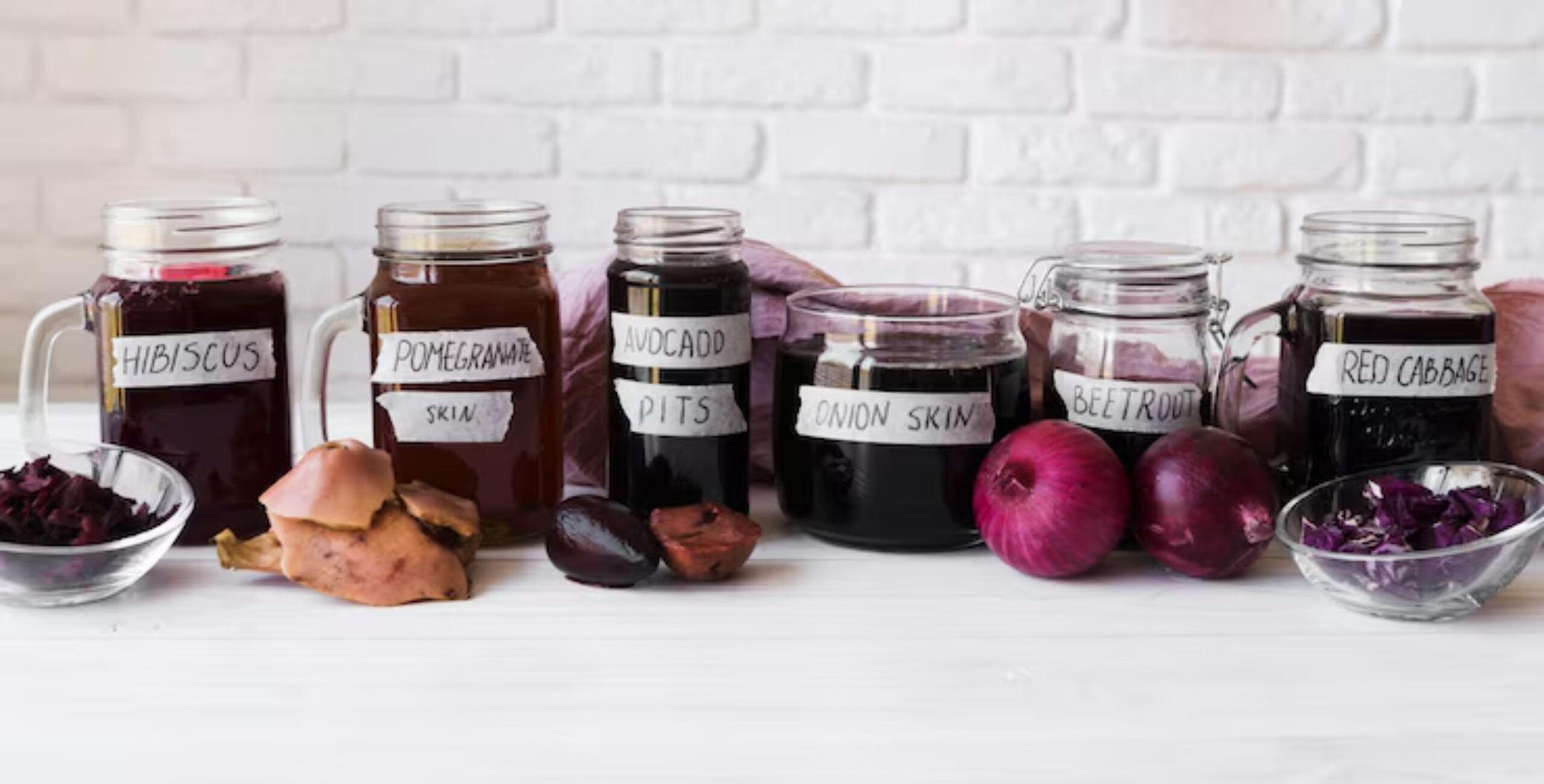 Jars of natural dye from various ingredients, labeled hibiscus, pomegranate, avocado pits, onion skin, beetroot, and red cabbage, on a table.