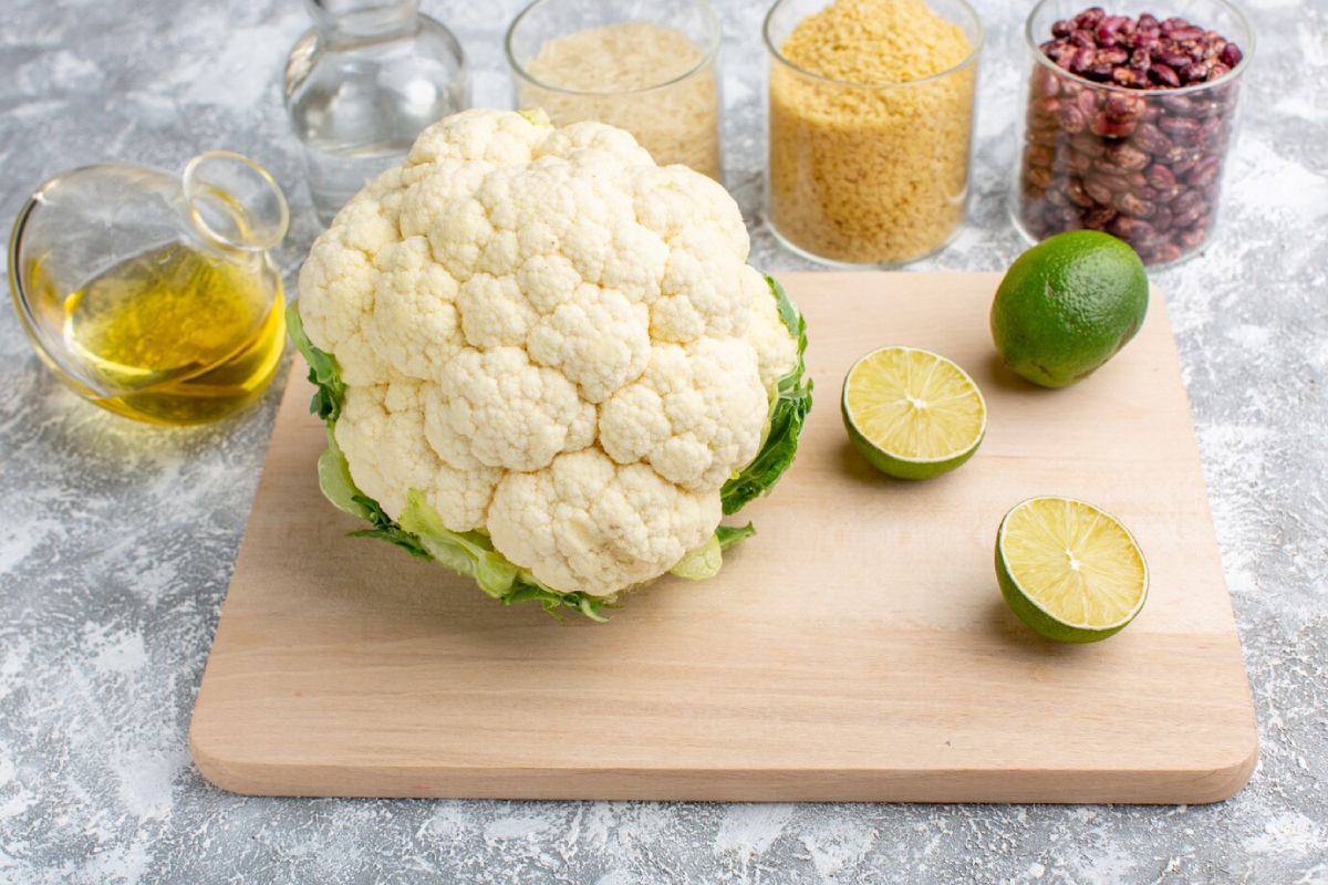 A fresh cauliflower sits on a wooden cutting board with lime halves, olive oil, and jars of grains and beans in the background.
