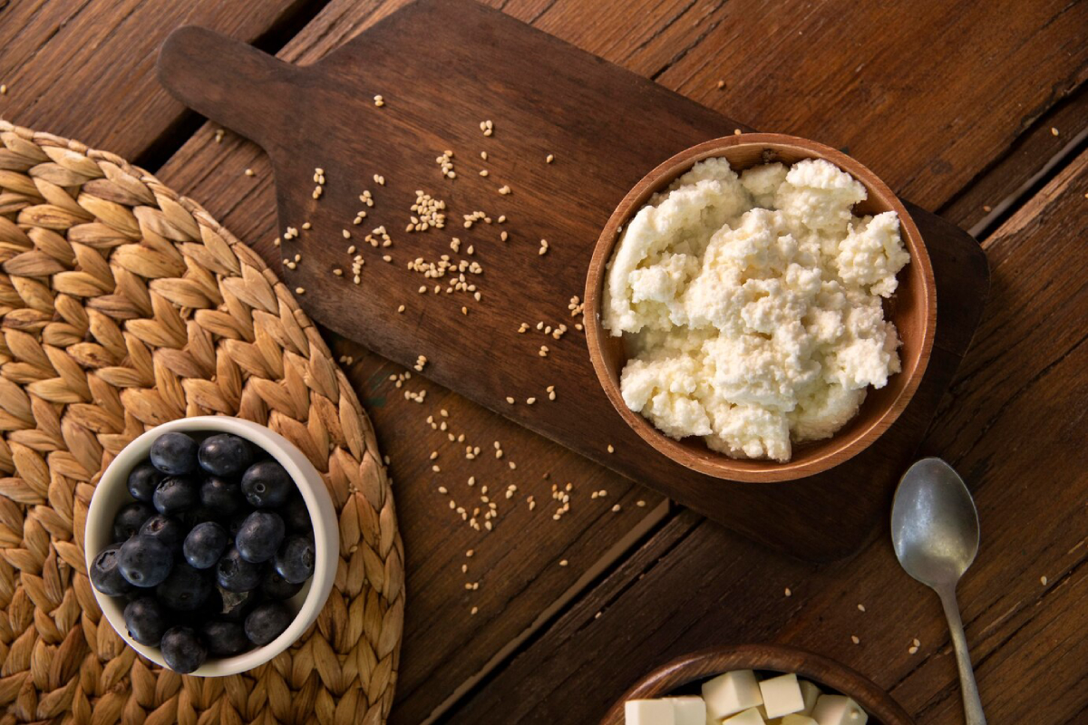 A bowl of ricotta cheese sits on a wooden board alongside a cup of blueberries and cubed cheese, with scattered sesame seeds around.