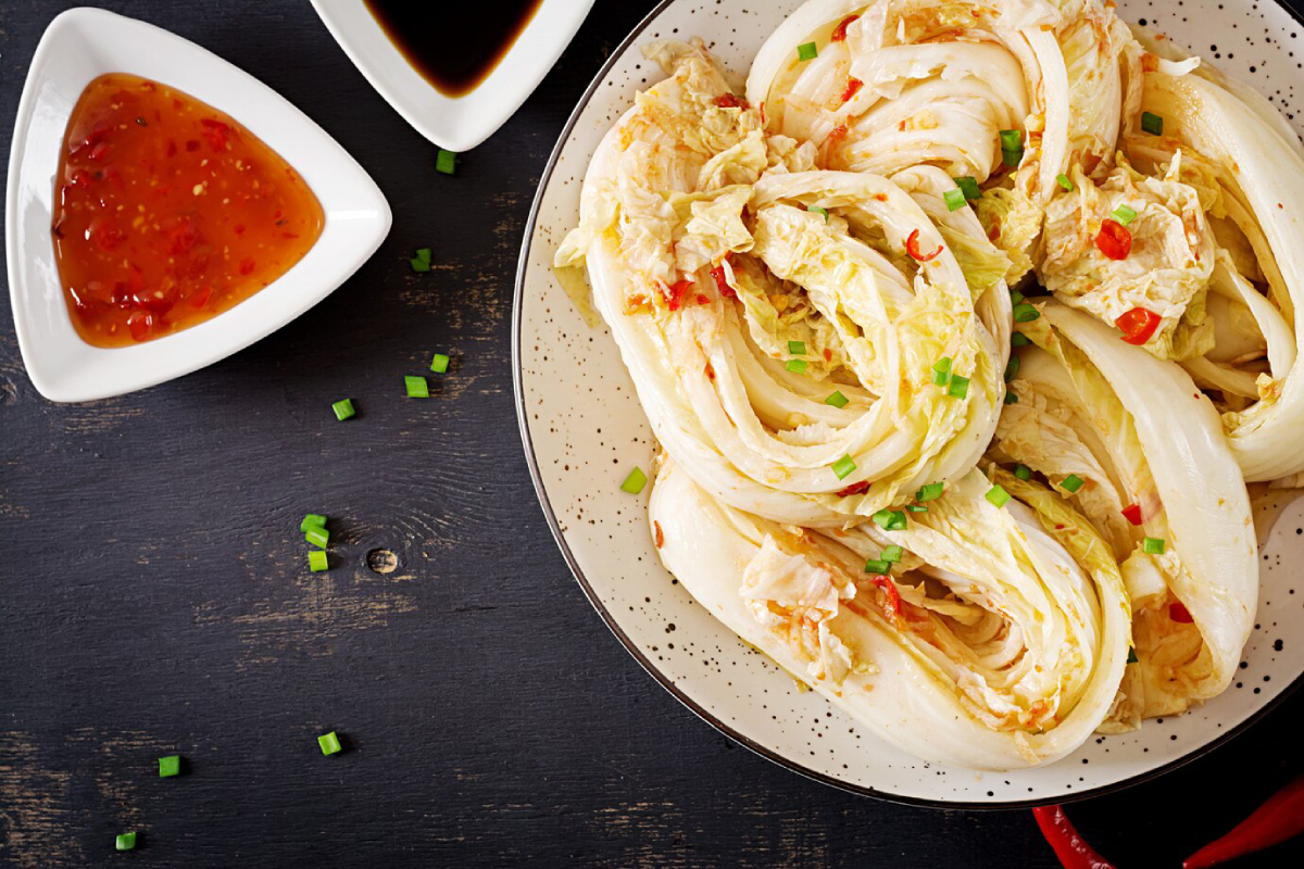 A plate of twisted napa cabbage garnished with chopped green onions and red chili, accompanied by dipping sauces on a dark wooden surface.