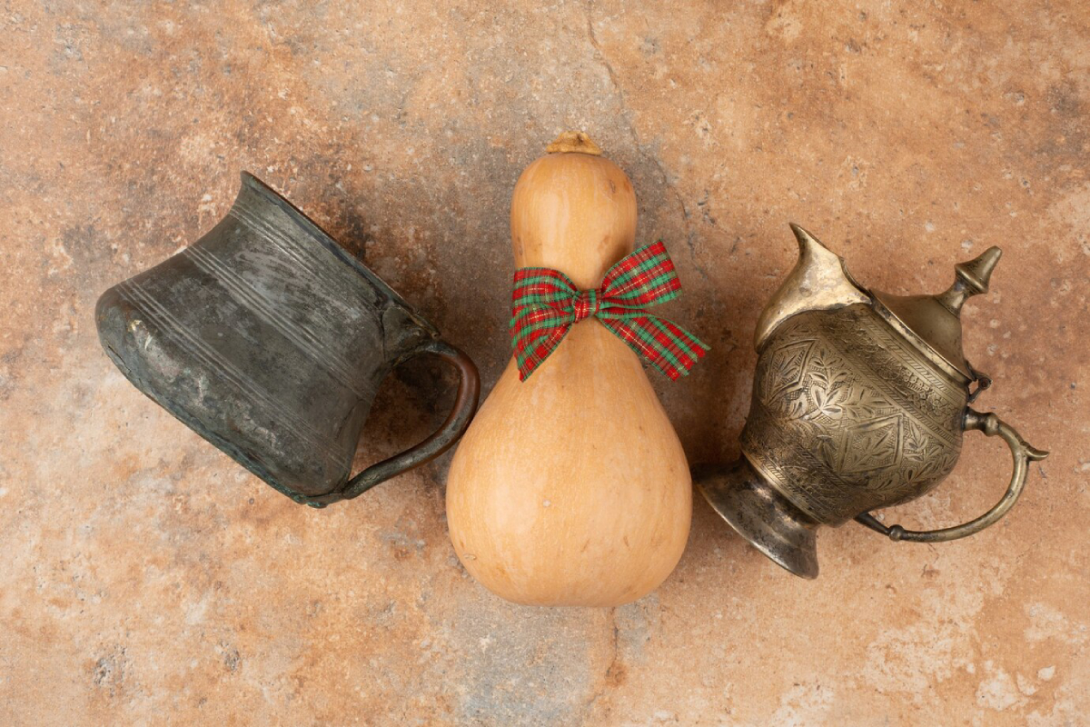 A rustic scene featuring a vintage metal cup, a gourd with a festive plaid bow, and an ornate metal teapot on a textured surface.