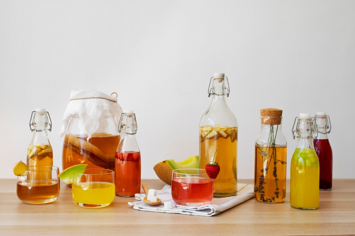 A variety of colorful beverages in glass bottles and glasses, featuring fruits, herbs, and a wooden spoon of sugar on a table.
