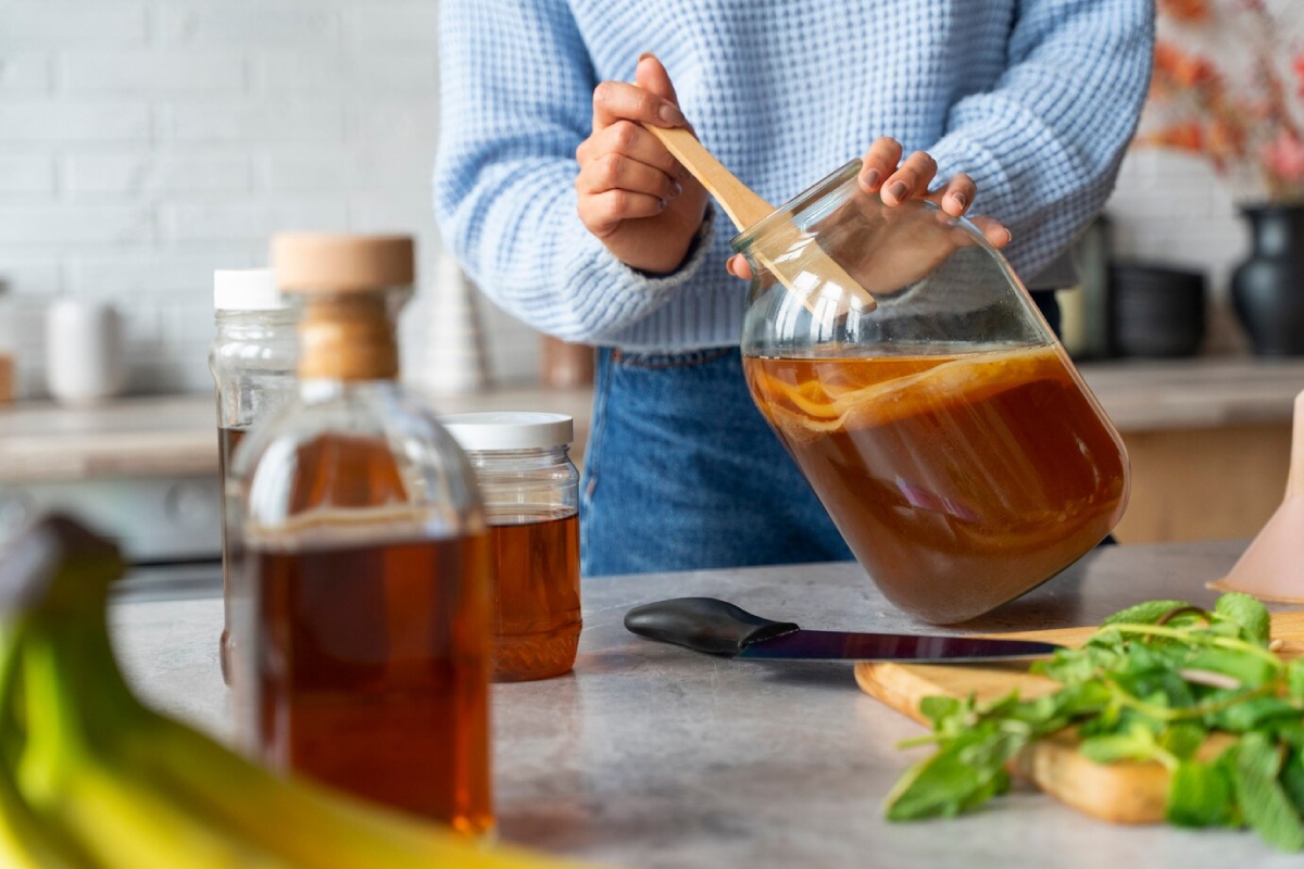 A person stirs a large glass jar of liquid with a wooden spoon, surrounded by bottles, bananas, and fresh mint on a kitchen countertop.